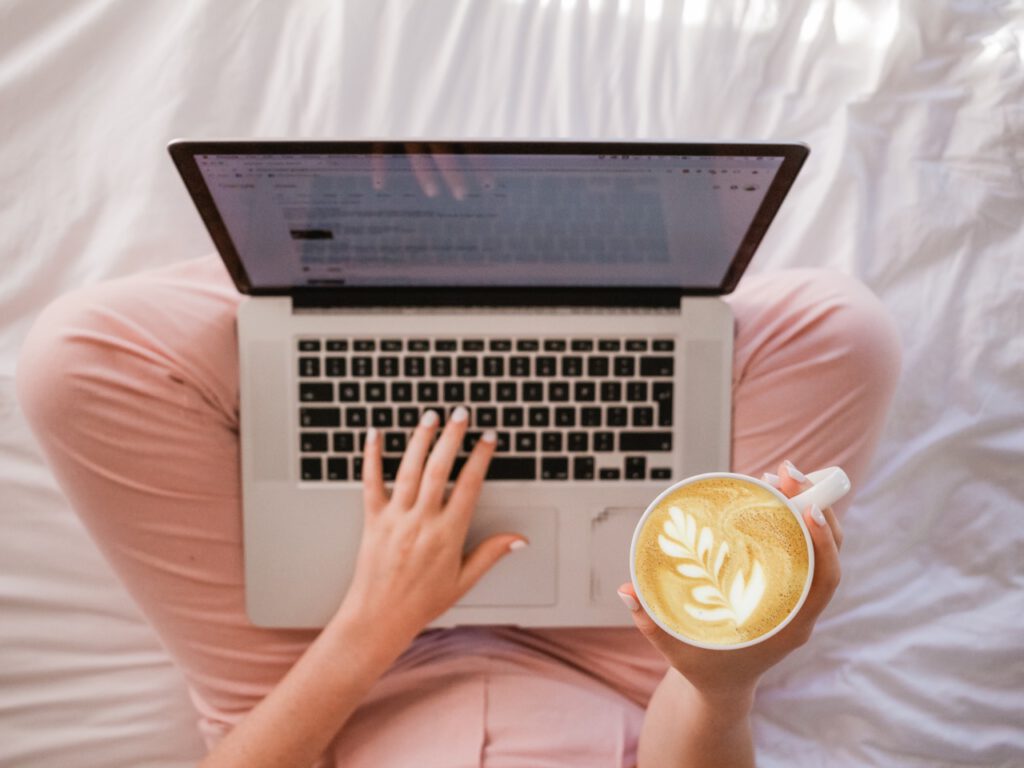 Een Macbook met een linker vrouwenhand die typt en de rechter die een cappuccino vasthoudt 