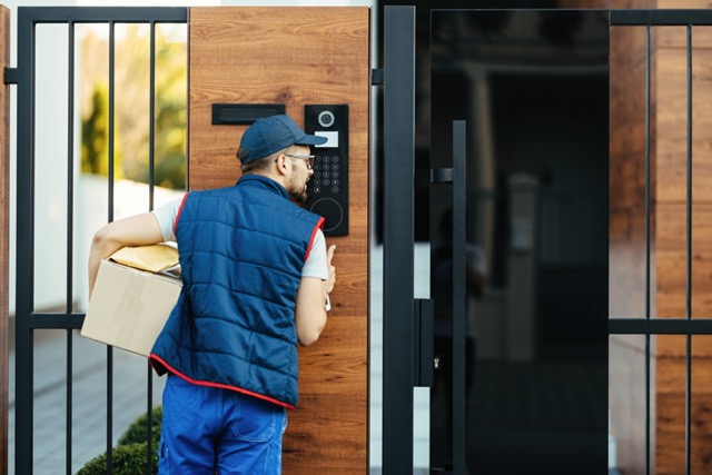 Back view of a deliverer ringing on intercom at gate of a customer's house while delivering packages.